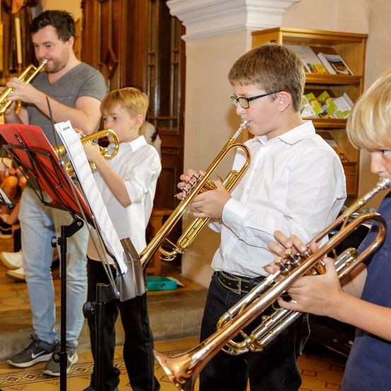 Ein Mann und drei Jungen spielen Trompeten in einem Raum mit einem Holzregal. Der Junge auf der rechten Seite trägt ein blaues Hemd.
