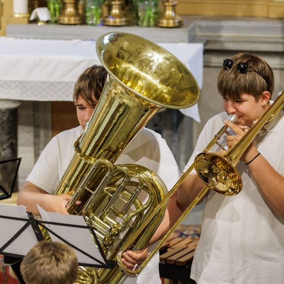 Zwei Jungen spielen Blechblasinstrumente in einer Kirche. Einer spielt eine Posaune und der andere eine Tuba. Ein dritter Junge beobachtet sie.