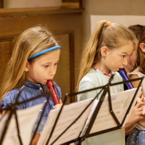 Zwei junge Mädchen spielen Blockflöten und lesen Musik von einem Notenständer. Ein Mädchen hat ein blaues Stirnband. Sie befinden sich wahrscheinlich in einem Klassenzimmer oder Musikraum.