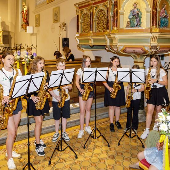 Eine Gruppe junger Musiker, wahrscheinlich Schüler, spielt Saxophone in einer Kirche. Der Raum ist mit Blumen und religiösen Statuen geschmückt.