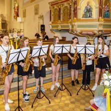 Eine Gruppe junger Musiker, wahrscheinlich Schüler, spielt Saxophone in einer Kirche. Der Raum ist mit Blumen und religiösen Statuen geschmückt.