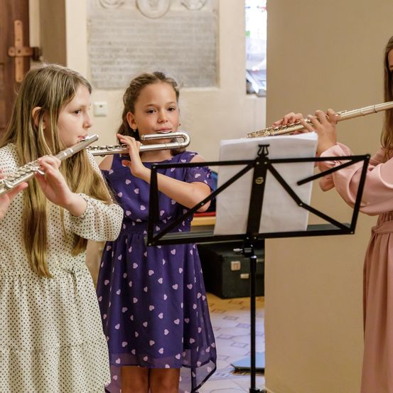Drei Mädchen spielen Flöten in einem Raum. Sie stehen um einen Notenständer mit einem Blatt Papier herum. Das Mädchen links trägt ein Punktekleid. Das Mädchen in der Mitte trägt ein lila Kleid mit Herzmustern. Das Mädchen rechts trägt ein rosa Kleid. Hinter ihnen ist eine Holztür.