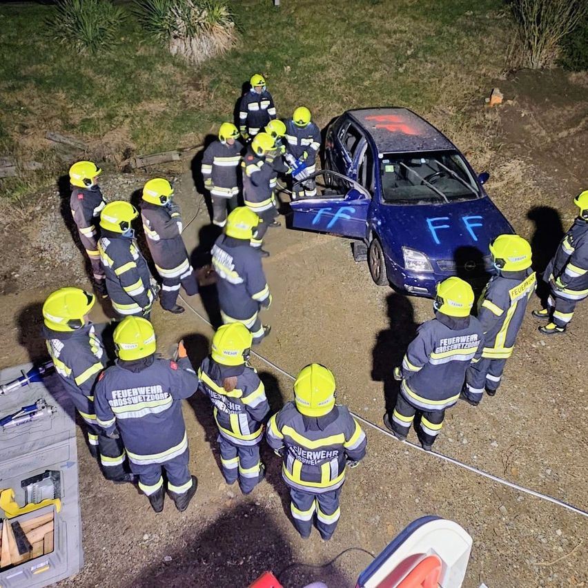 Eine Gruppe von Feuerwehrleuten in gelben Helmen und Uniformen steht um ein blaues Auto herum, auf dem 'FF' mit Sprühfarbe bemalt ist. Sie stehen auf einem Erdweg.