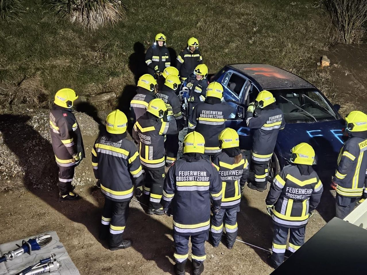 Eine Gruppe von Feuerwehrleuten in gelben Helmen und Uniformen versammelt sich um ein beschädigtes Auto auf einem Hang.