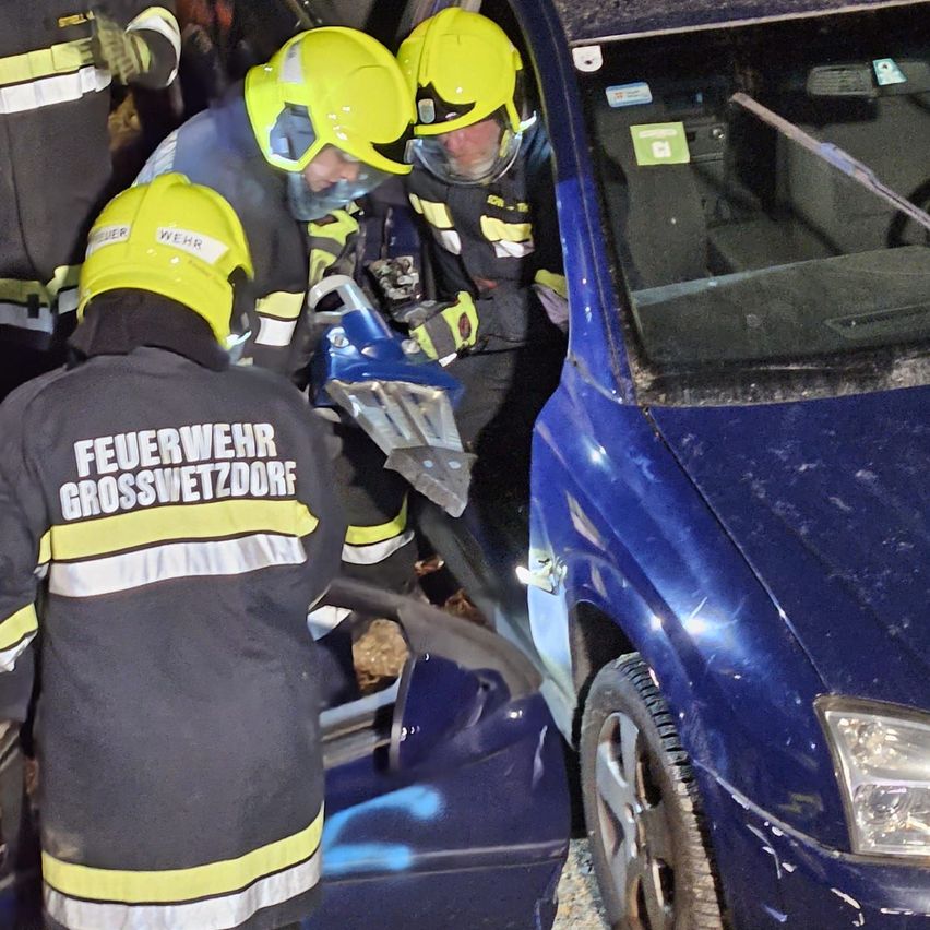Feuerwehrleute in gelben Helmen arbeiten an einem blauen Auto, möglicherweise nach einem Unfall. Die Autotür steht offen, und die Windschutzscheibe zeigt Reflexionen.