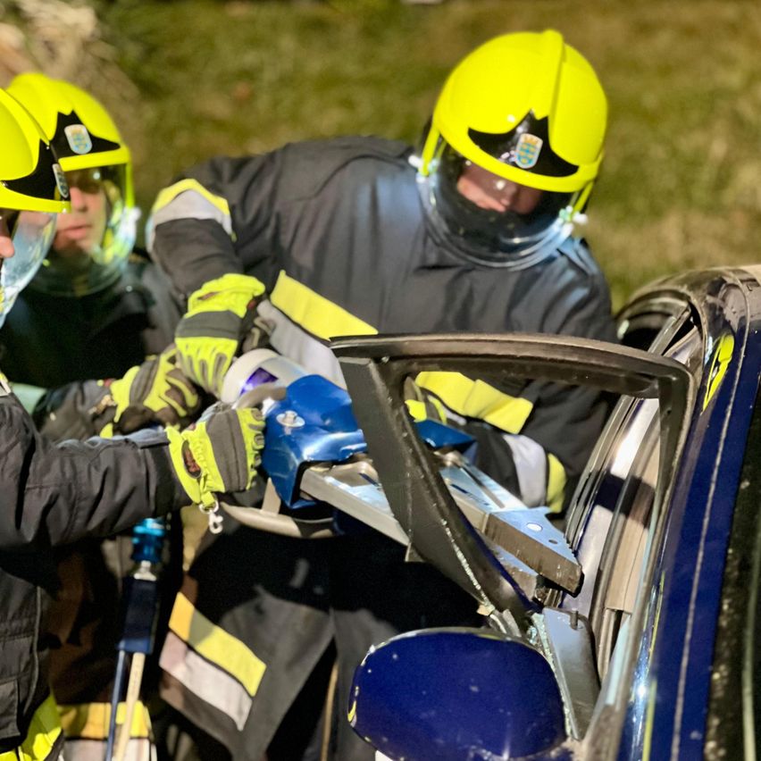 Drei Feuerwehrleute in Schutzausrüstung arbeiten bei Nacht daran, eine beschädigte Autotür zu öffnen. Sie benutzen ein Werkzeug, um die Tür aufzuhebeln.