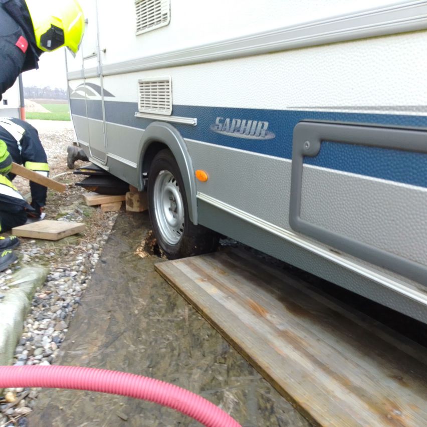 Feuerwehrleute arbeiten daran, einen Wohnwagen zu befreien, der auf einem Kiesweg feststeckt. Der Reifen des Wohnwagens wird mit Holzklötzen angehoben, und ein roter Schlauch liegt in der Nähe.