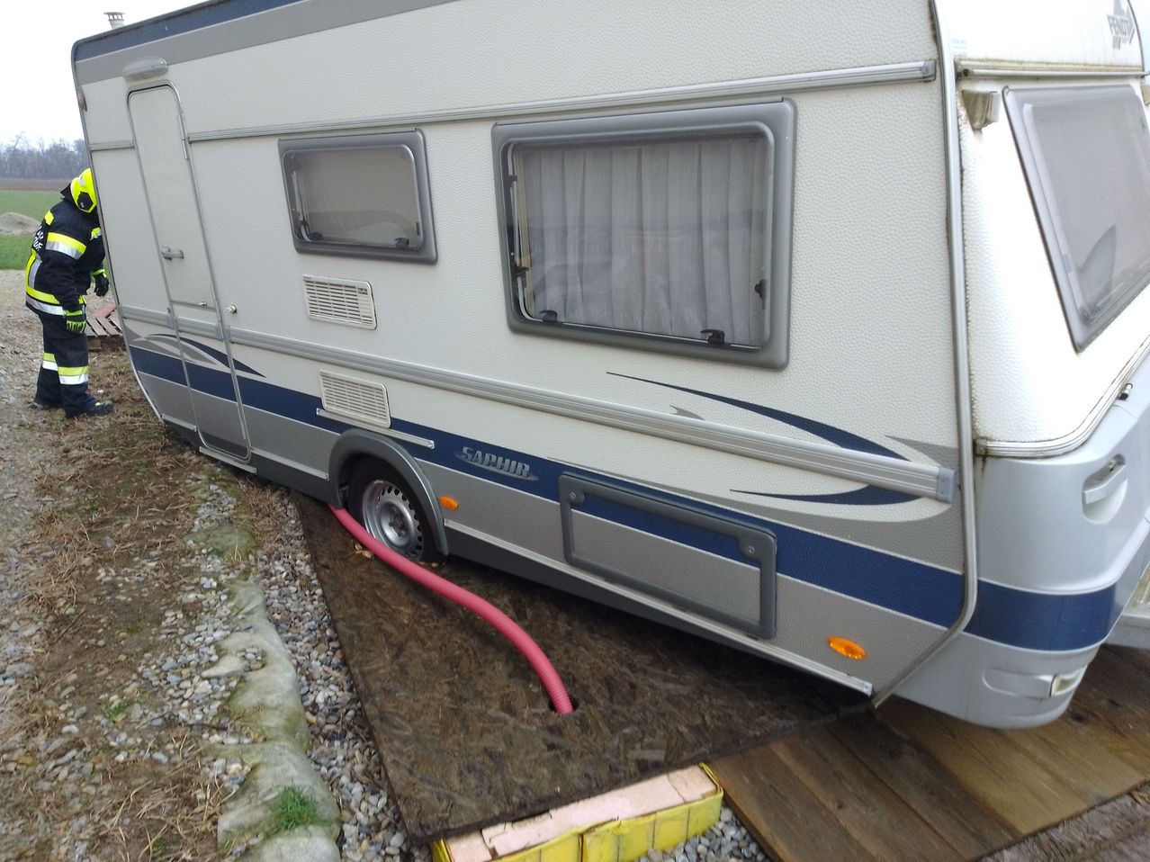 Ein weißer und blauer Wohnwagen steht auf einem Schotterboden, an dem ein roter Schlauch angeschlossen ist.