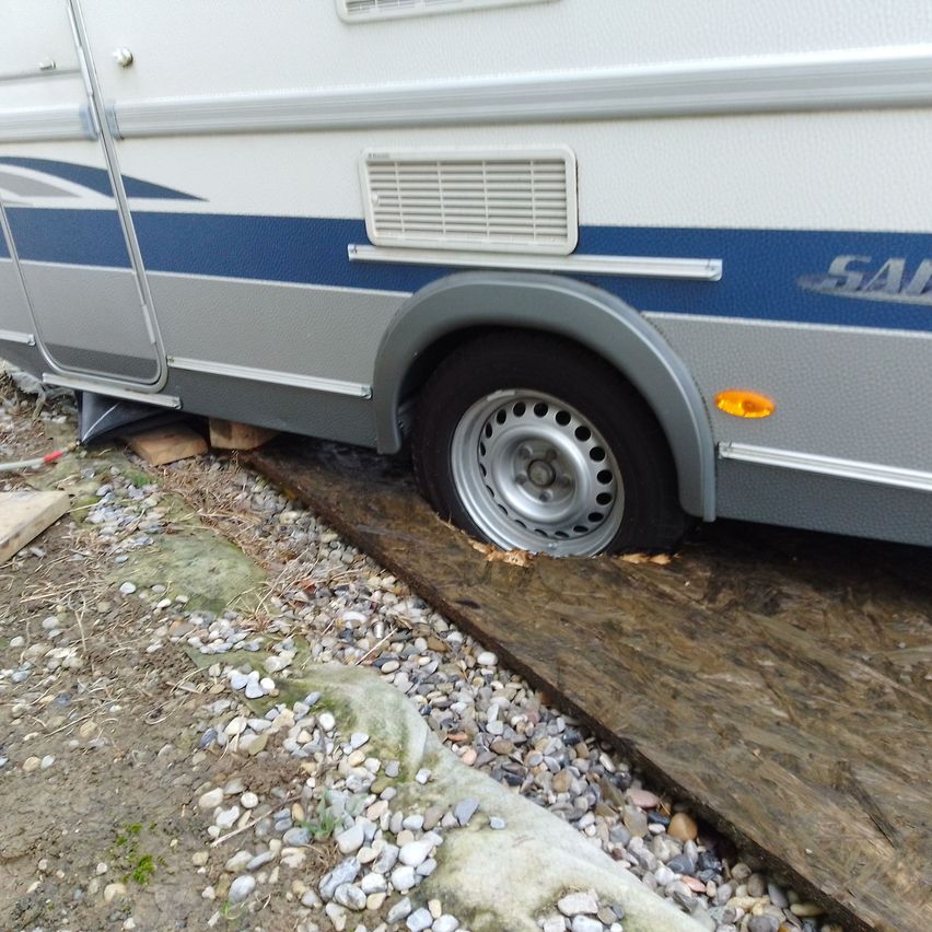 Ein weißer und blauer Wohnwagen mit der Marke SAI steht auf einer Kiesoberfläche mit einem platten Reifen.
