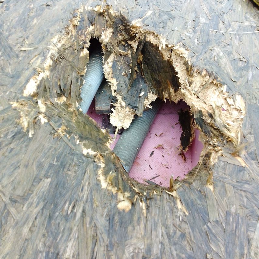 Eine Nahaufnahme eines Lochs in einem Dach mit freiliegendem Holz und Isolierung, wobei zwei Rohre durch das Loch ragen.