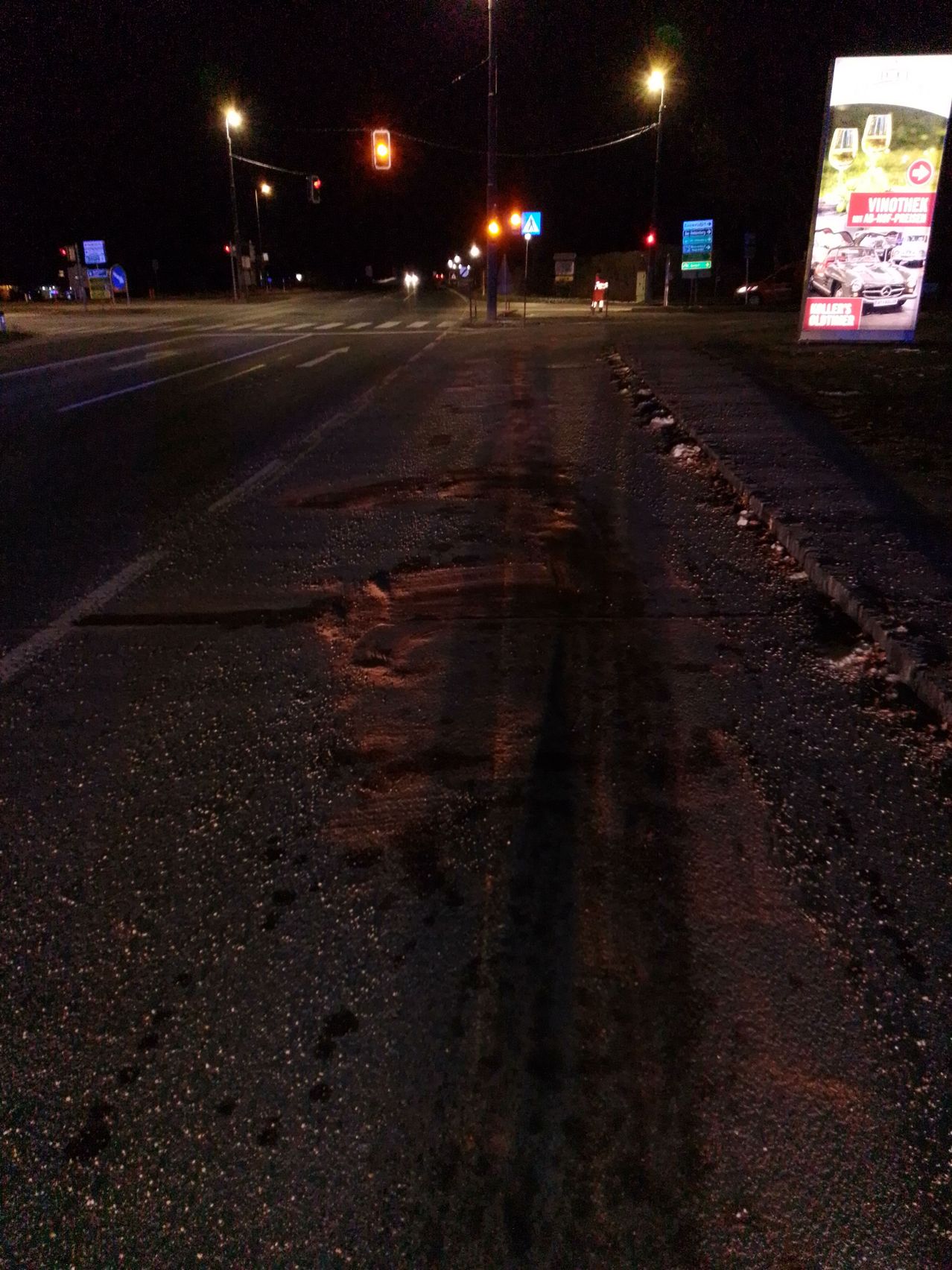 Ein Nachtblick einer leeren Straße mit Schlaglöchern und Ampeln. Eine Person läuft auf dem Bürgersteig. Eine große Werbung befindet sich am Straßenrand.