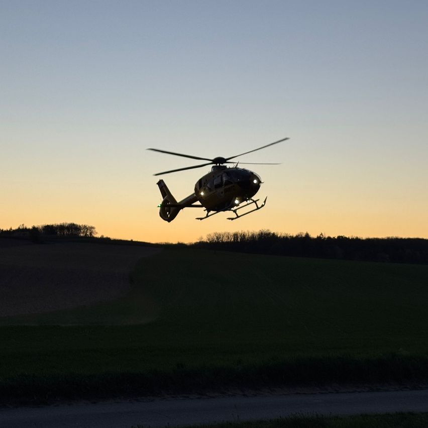 Ein Hubschrauber schwebt bei Dämmerung über einem Feld, das Fahrwerk ist ausgefahren.