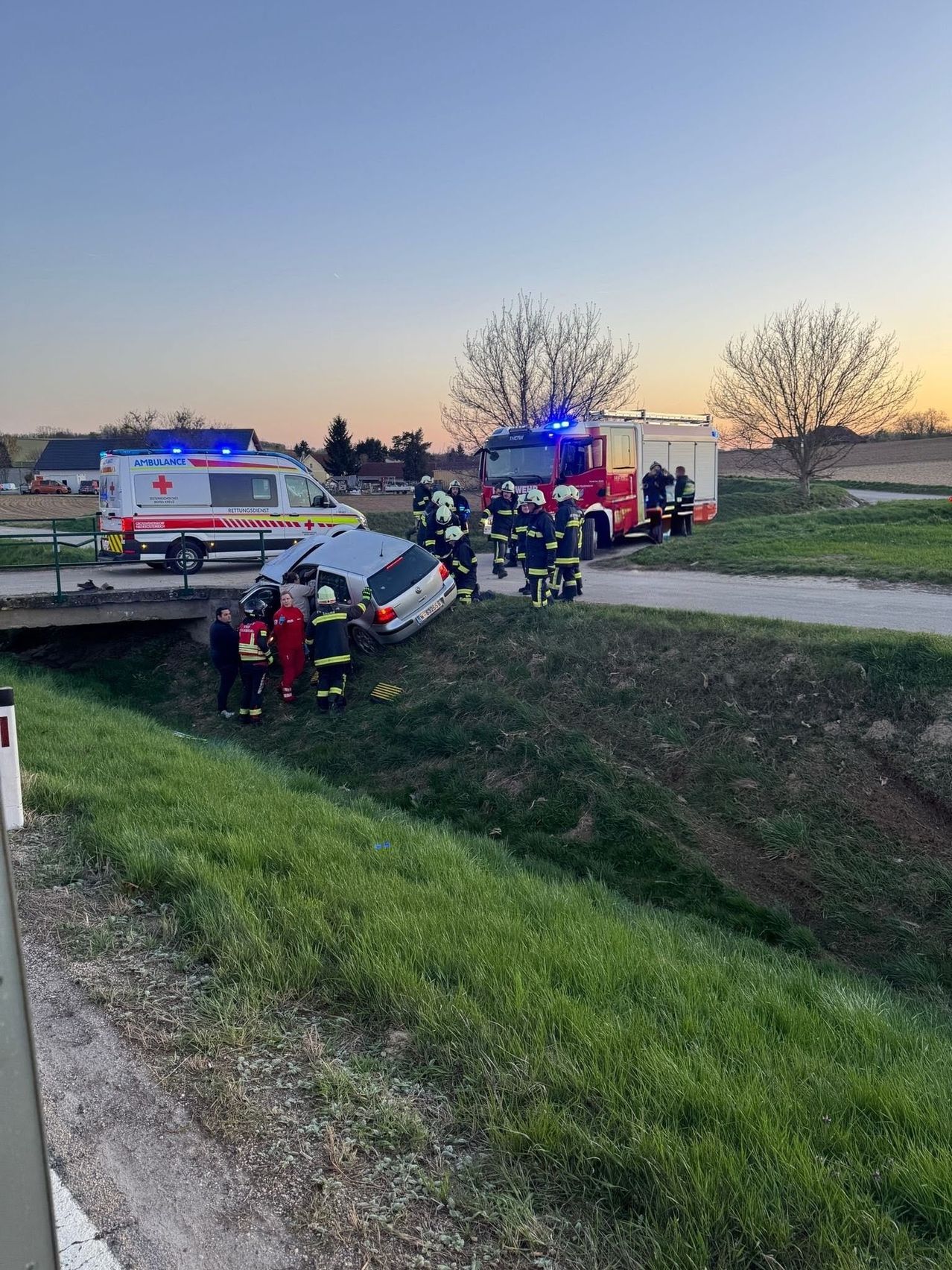 Ein umgestürztes Auto wird von Rettungskräften versorgt. Das Fahrzeug liegt auf der Seite an einem Grashang. In der Nähe stehen Feuerwehrleute und Sanitäter.