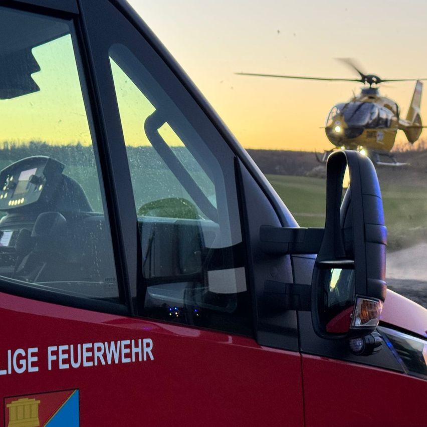 Ein roter Feuerwehrwagen ist in der Nähe eines Grasfeldes geparkt. Das Wort 'FEUERWEHR' steht auf der Seite. Ein gelber Hubschrauber fliegt über dem Feld.