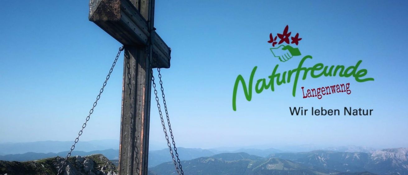 In einer Berglandschaft steht ein hölzernes Kreuz mit Ketten prominent. Ein Logo mit den Worten 'Naturfreunde Lang' und 'Wir le' ist überlagert. Der Himmel ist klar und blau.