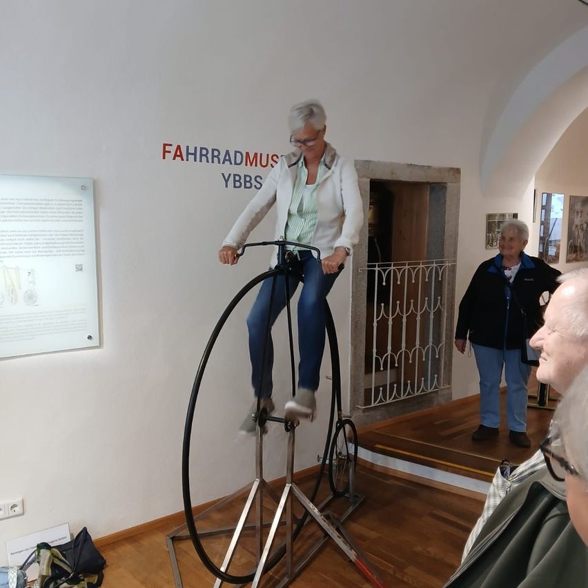 Eine ältere Frau mit Brille fährt ein altes Fahrrad in einem Museum, während zwei andere Personen zusehen. Die Wand ist mit 'FAHRRADMUS YBBS' beschriftet.