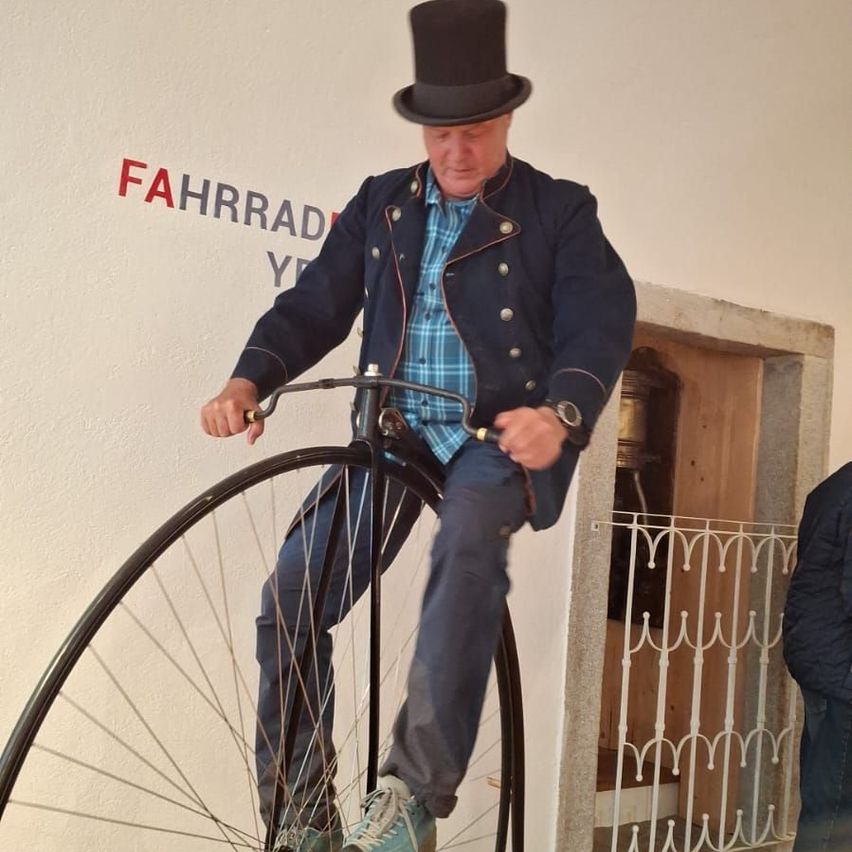 Ein Mann in einem hohen Hut fährt ein altes Penny-Farthing-Fahrrad. Er steht auf den Pedalen, und ein Schild mit der Aufschrift 'FAHRRAD' ist an der Wand zu sehen.