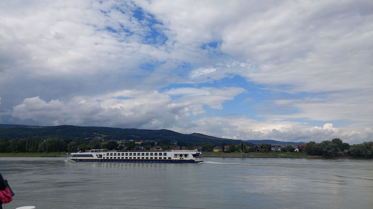 Ein großes weißes Boot fährt auf dem Fluss unter einem blauen Himmel mit verstreuten Wolken. Die Berge sind im Hintergrund.