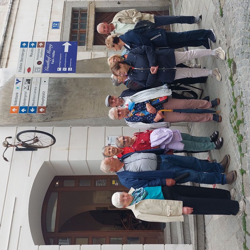 Eine Gruppe von Menschen steht auf einem Steinweg, einige halten Regenschirme. Sie sind in der Nähe eines Gebäudes mit einem gewölbten Eingang. Ein Fahrrad hängt an der Wand.