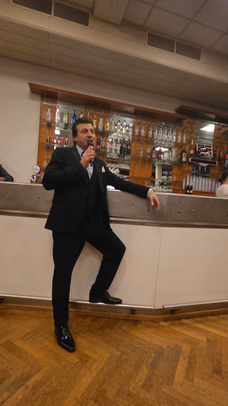 Ein Mann in einem schwarzen Anzug steht auf einer Theke und hält ein Mikrofon. Dahinter ist eine Bar mit Regalen voller Likörflaschen zu sehen.