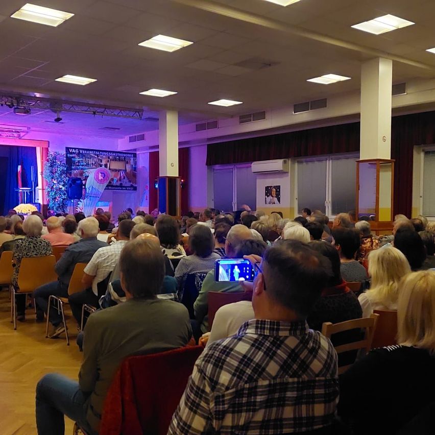 Eine Menschenmenge sitzt auf Stühlen vor einer Bühne. Eine Person macht ein Foto mit einem Smartphone. Die Bühne hat einen Bildschirm und ein Banner mit Text. Die Decke hat angebrachte Lampen.