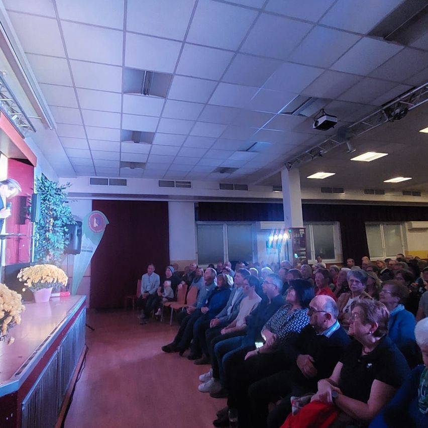 Ein Saal mit Bühne hat ein Publikum, das in Reihen sitzt. Die Bühne hat eine Pflanze und einen Lautsprecher. Das Publikum schaut sich eine Aufführung an. Die Decke hat Lichter und einen Projektor.