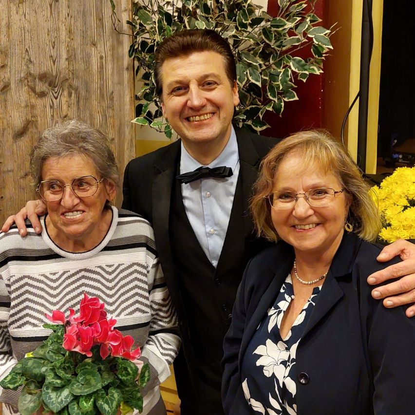 Ein Mann in einem Anzug steht zwischen zwei Frauen, alle lächeln. Die linke Hand des Mannes liegt auf der Schulter der Frau rechts von ihm. Die Frau links hält einen Strauß pinker Blumen. Eine grüne Pflanze mit gelben Blüten steht hinter ihnen.