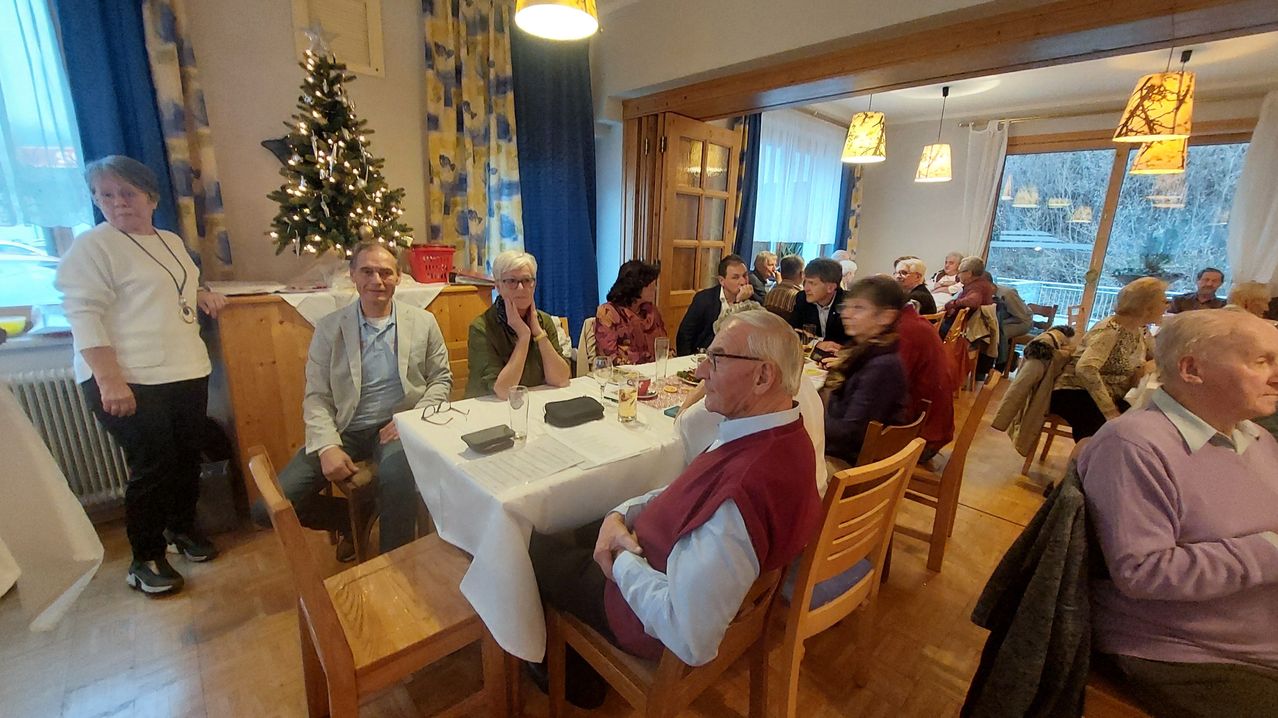Eine Gruppe von Menschen sitzt in einem Esszimmer mit Weihnachtsdekoration. Einige unterhalten sich, während andere auf ihre Mahlzeiten konzentriert sind.