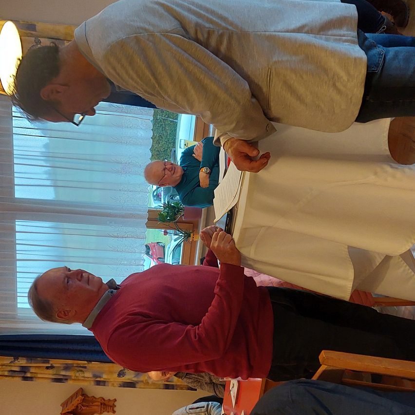 Ein Mann schaut einen anderen Mann an, der einen Stift und Papier hält. Hinter ihnen steht ein Schreibtisch mit einer Pflanze und einem Fenster mit Jalousien. Ein dritter Mann sitzt und schaut etwas an.