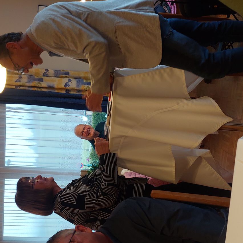 Eine Gruppe von Menschen versammelt sich in einem Raum. Ein Mann beugt sich über einen Tisch mit einem weißen Tuch. Eine Frau mit Brille sitzt in der Nähe. Ein anderer Mann steht hinter dem Tisch. Sie scheinen ein Gespräch zu führen.