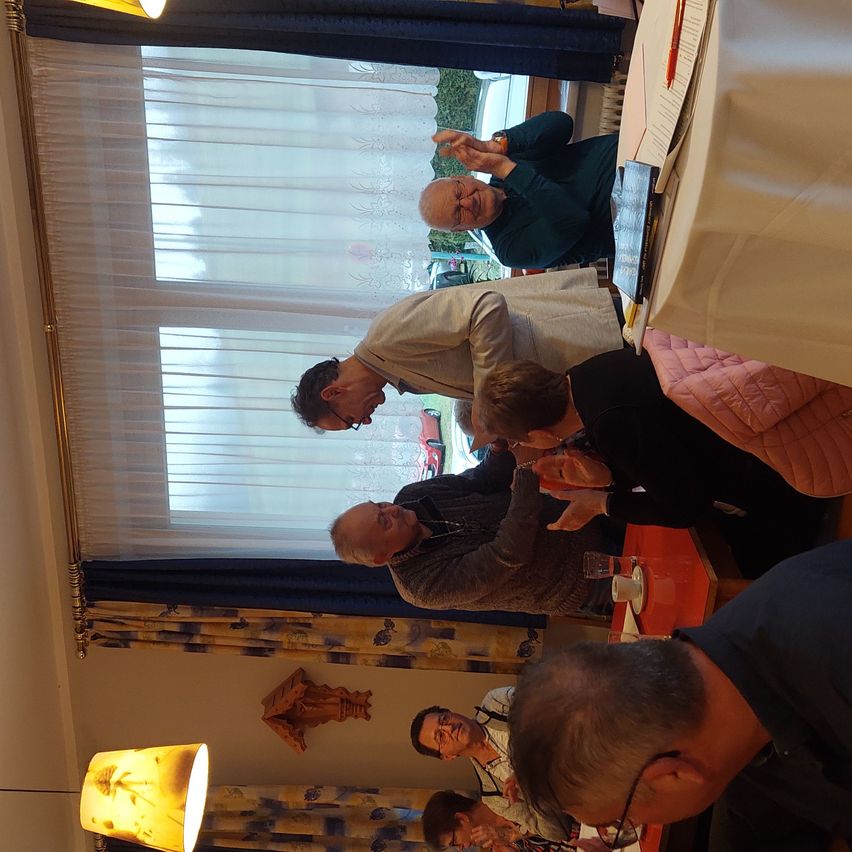 Eine Gruppe von Menschen versammelt sich in einem Raum, anscheinend in einer Besprechung. Einige stehen, andere sitzen. Hinter ihnen befindet sich ein Fenster mit Vorhängen.