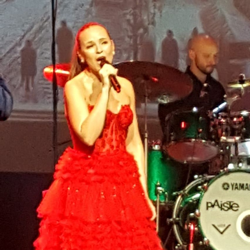 Eine Frau in einem roten Kleid singt in ein Mikrofon auf der Bühne, mit einem Schlagzeug hinter ihr. Ein Mann spielt das Schlagzeug.