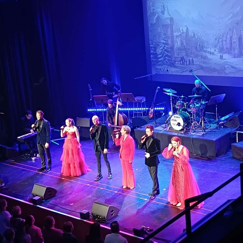 Eine Aufführung auf einer Bühne mit einer Band und Sängern, eine Frau in einem roten Kleid, eine andere in Pink. Ein Hintergrund zeigt eine verschneite Szene.