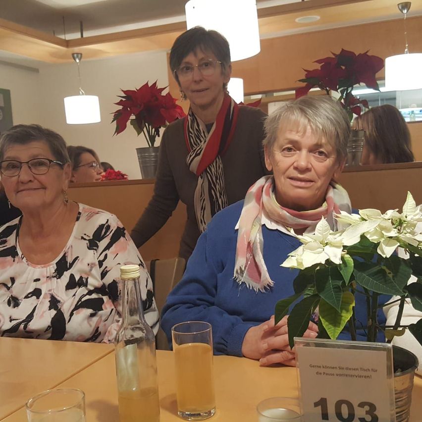 Drei ältere Frauen sitzen an einem Tisch, eine mit einer Flasche, Gläsern mit Saft und einer Pflanze. Eine andere trägt eine Schals. Hinter ihnen steht eine Frau in der Nähe eines Tisches mit roten Blumen. Der Tisch hat ein Schild mit der Nummer 103.