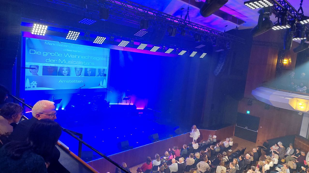 Ein Weihnachts-Musical-Event mit einer Bühne, auf der ein großer Bildschirm Bilder von Menschen zeigt. Eine Frau steht auf der Bühne, während das Publikum zuschaut. Die Bühne ist mit blauen und lila Lichtern beleuchtet.