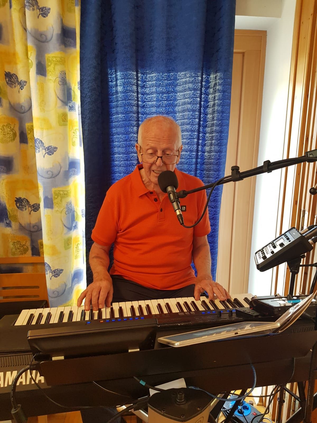 An older man wearing an orange shirt plays a keyboard while smiling into a microphone, with blue curtains in the background.