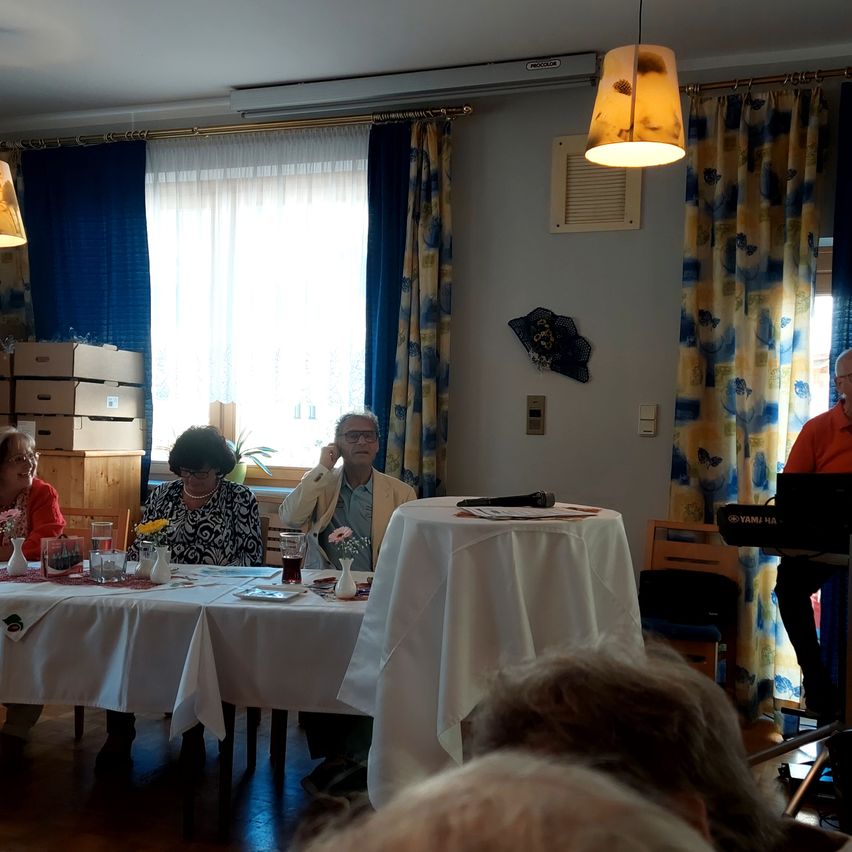 A meeting in progress. Four individuals are seated around a table. A man in the middle holds a microphone. Behind them is a window with curtains.