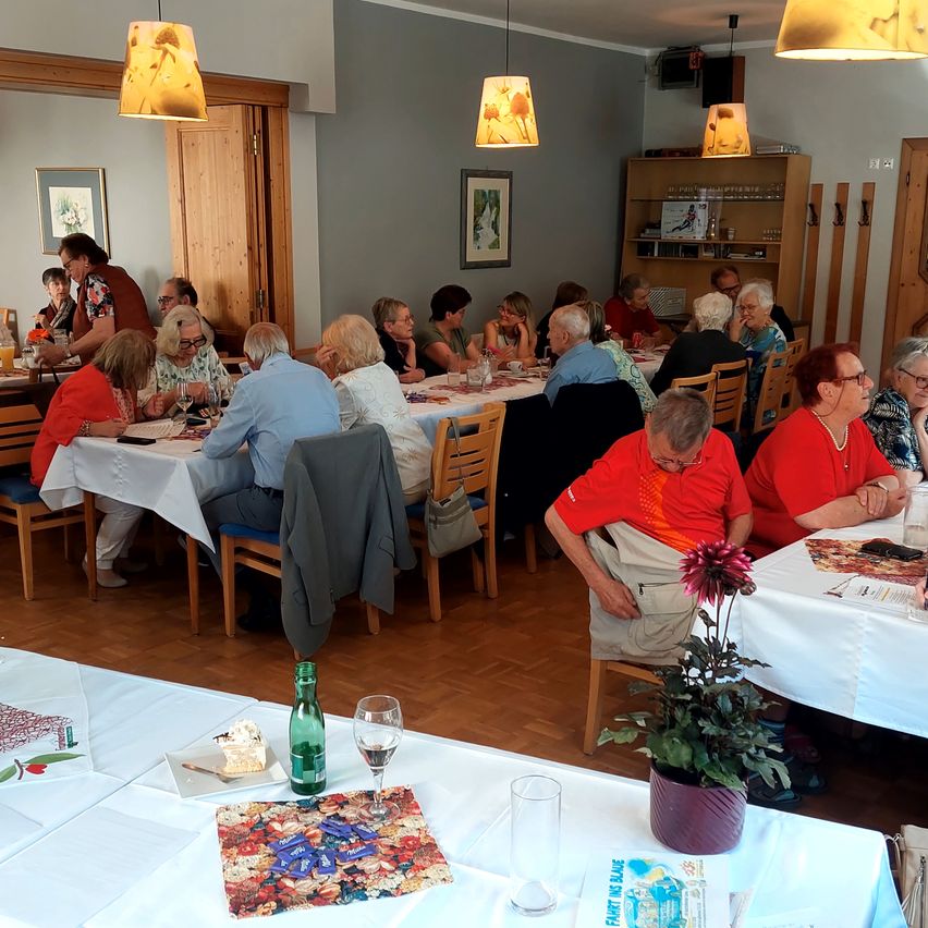 A group of elderly people sit around tables in a room, some eating and some talking. Tables are covered with white cloths, and there are flowers and glasses on some tables.