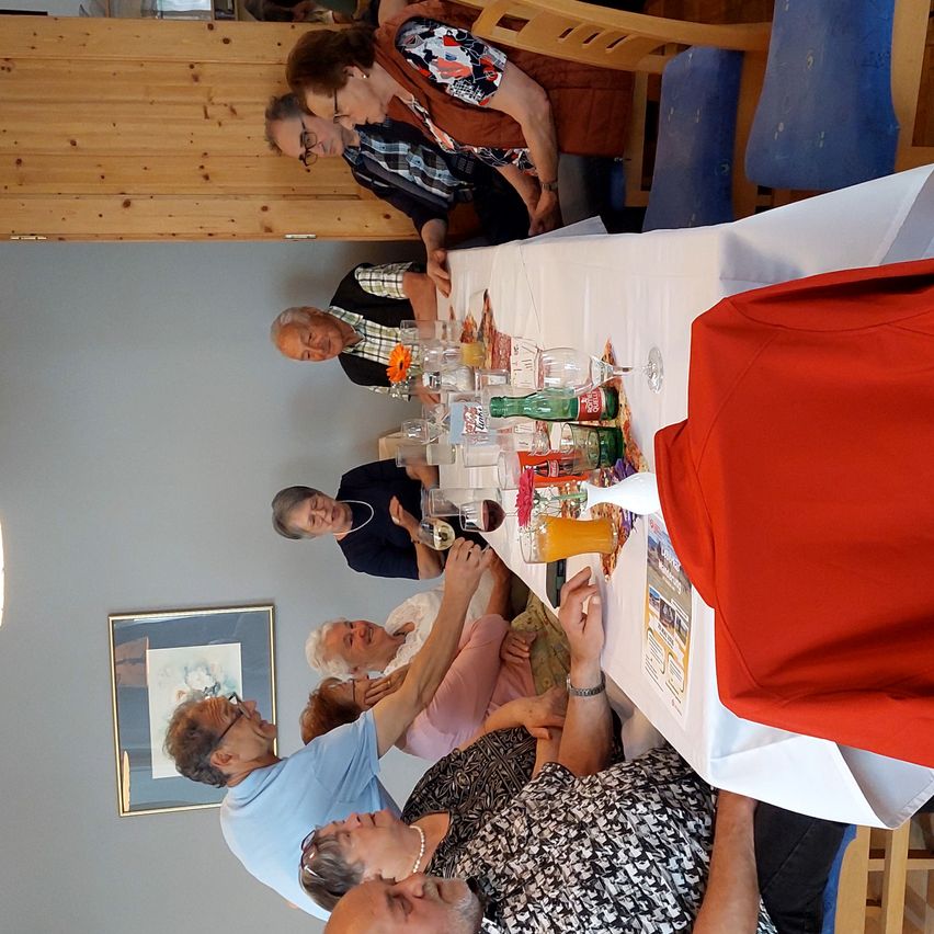 Several people sit around a table, possibly at a family gathering. The table is laden with various drinks and snacks. Some people wear glasses. A framed picture is mounted on the wall.