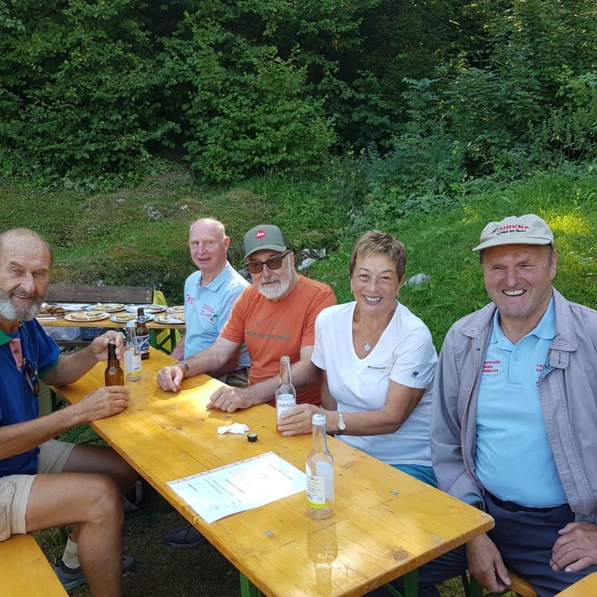 Fünf Personen sitzen um einen Holztisch in einem Park, lächeln und posieren für ein Foto. Sie haben Flaschen und Teller auf dem Tisch. Der Hintergrund ist eine üppige grüne Landschaft mit Bäumen.