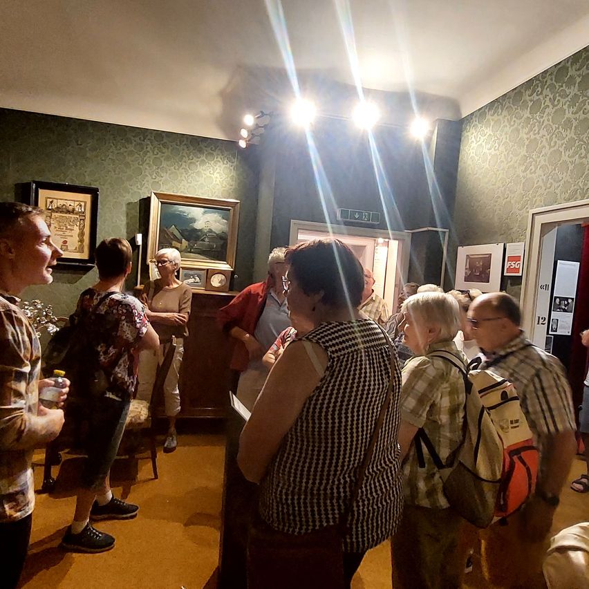 A group of people are standing in a room, possibly a museum, with one person holding a paper. The room has a wooden desk, a picture frame, and a lamp.