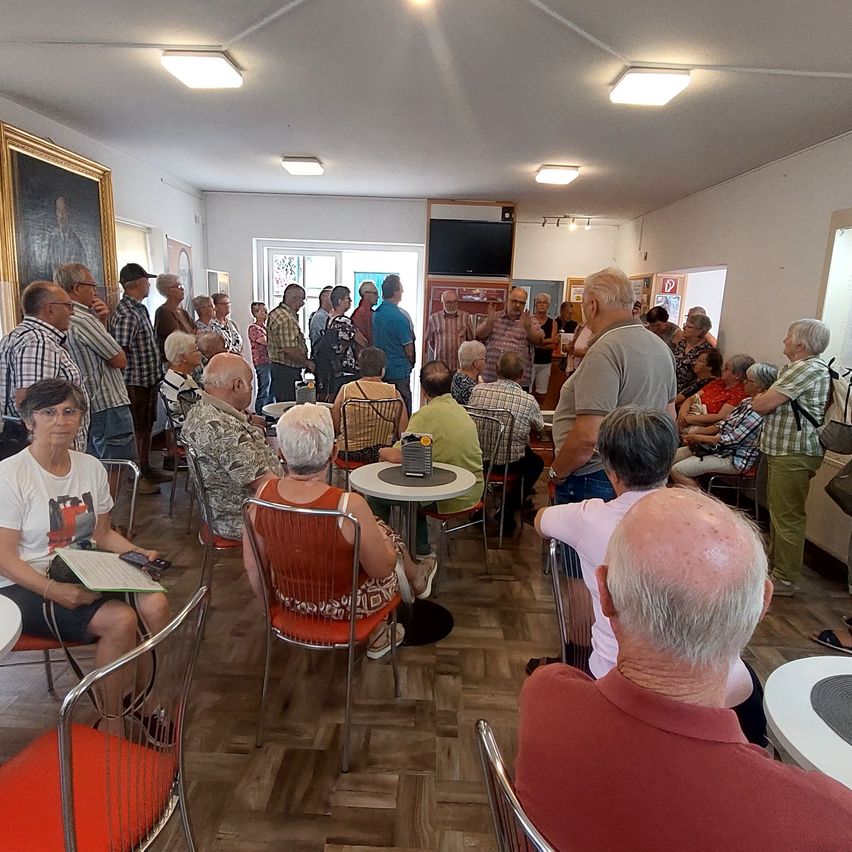 A group of elderly people are gathered in a room, some standing and some sitting on chairs. A woman in the front holds a book. A table with a cup and a monitor is in the center.