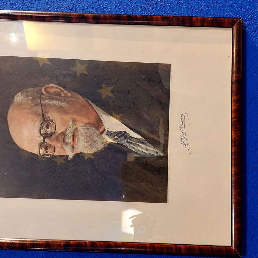 A framed portrait of an elderly man with a white beard and mustache wearing glasses and a suit. The portrait has a blue background with stars.