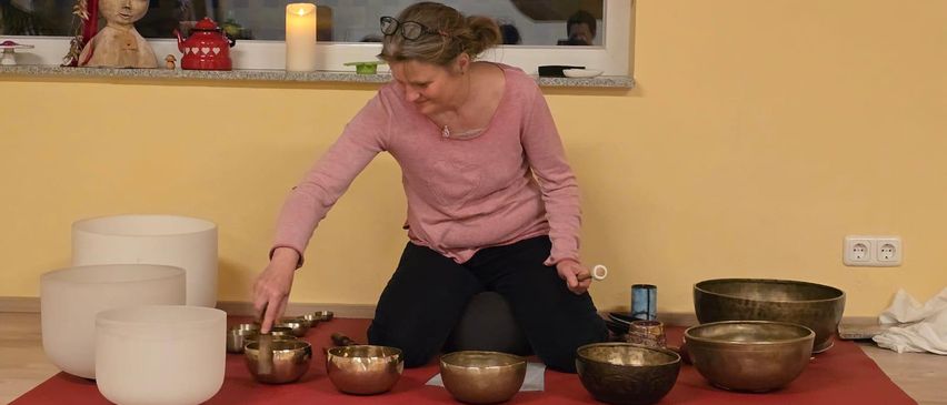 Eine Frau kniet auf einem roten Teppich und spielt mit tibetanischen Klangschalen. Sie trägt ein rosa Hemd und eine Brille. Vor ihr stehen mehrere Schalen auf dem Teppich und eine brennende Kerze auf einem Fenstersims hinter ihr.