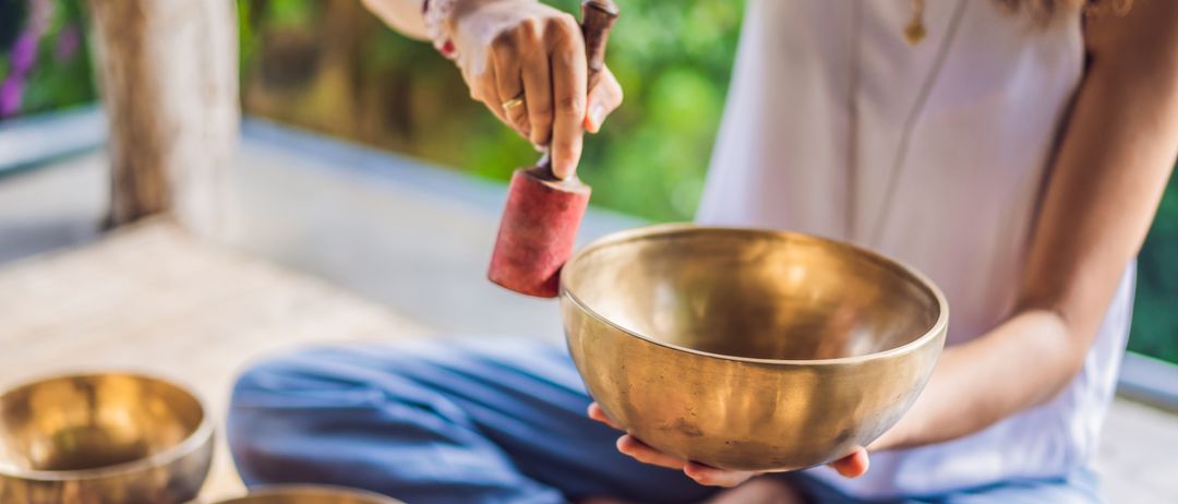 Eine Frau sitzt im Schneidersitz auf dem Boden und hält eine bronzene Klangschale. Sie schlägt sanft mit einem hölzernen Schlägel darauf.