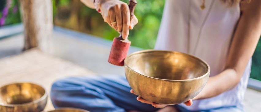 Eine Frau sitzt im Schneidersitz auf dem Boden und hält eine bronzene Klangschale. Sie schlägt sanft mit einem hölzernen Schlägel darauf.