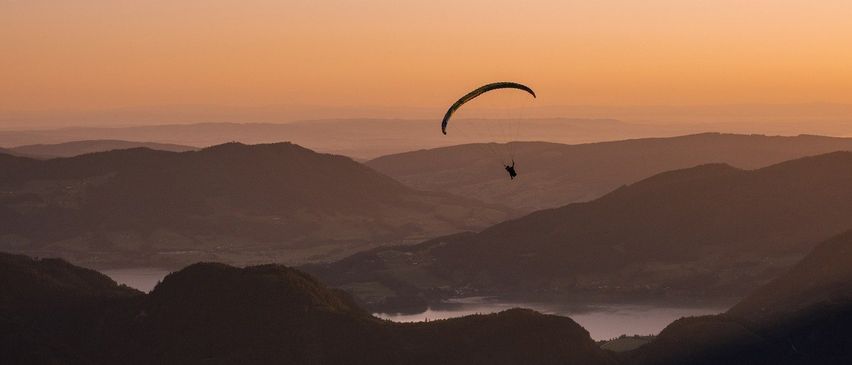 Eine Person paragliding über eine bergige Landschaft während eines Sonnenuntergangs, mit einem ruhigen See darunter.