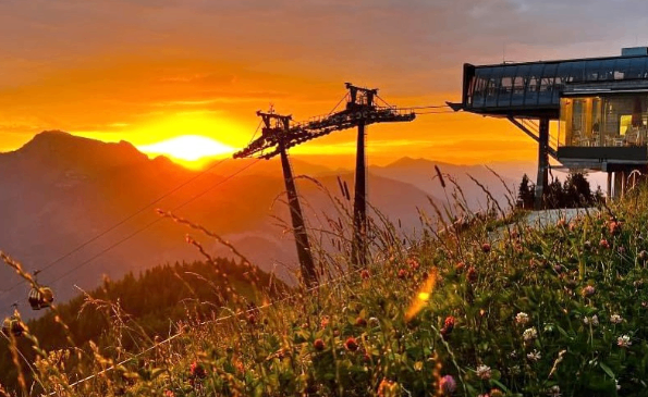 Sonnenuntergang über Bergen mit einer Seilbahnstation im Vordergrund. Der Himmel ist orange, und ein Gebäude ist im Hintergrund sichtbar.