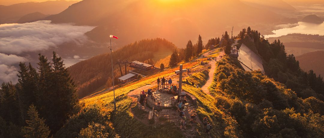 Ein Sonnenuntergang vom Berggipfel zeigt ein Kreuz und eine Gruppe von Menschen, die darum stehen. Der Himmel ist orange und pink, mit Bergen und einem See in der Ferne.