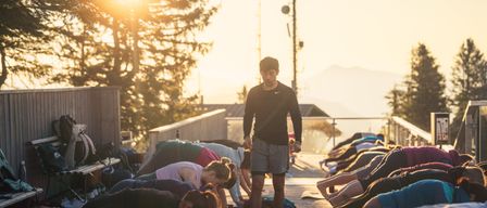 Eine Gruppe von Menschen praktiziert Yoga auf einer Terrasse bei Sonnenuntergang, ein Mann steht vor ihnen.